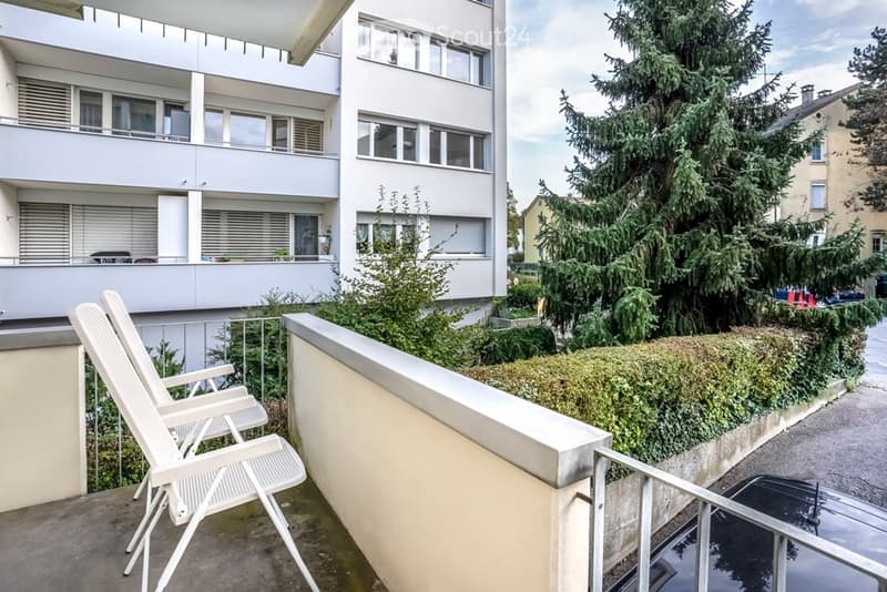 Balcony with white lounge chairs, metal railing, and a view of a modern apartment building and greenery.