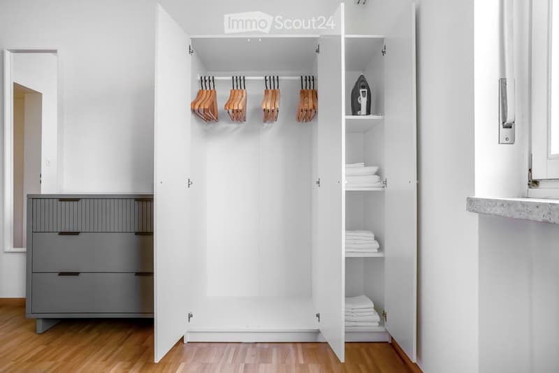 Modern, minimalist walk-in closet with white sliding doors, wooden floor, grey drawer unit, and neatly organized clothing and towels.