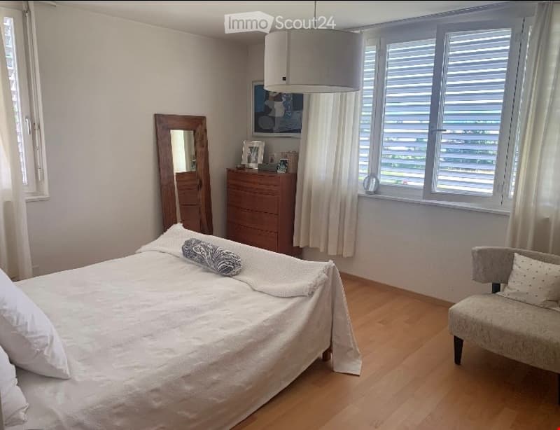 Bright bedroom with wooden flooring, a neatly made bed, a small armchair, a dresser with a mirror, and a picture frame on the wall.
