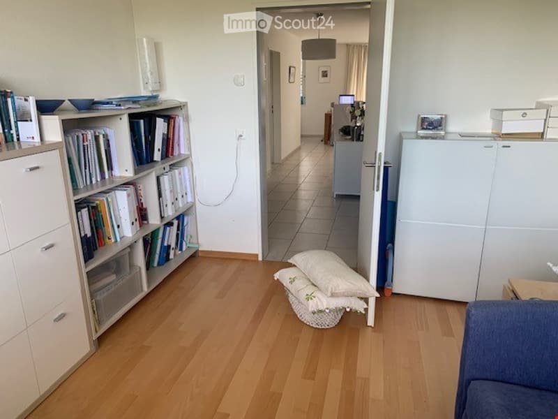 A modern, well-lit living room with light wooden flooring, a blue sofa, a white bookshelf filled with books, a small table with pillows, and a view into an adjacent room with a refrigerator and a table.