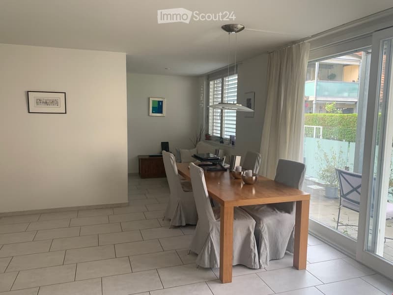 Dining room with a wooden dining table, six chairs, and a view to a balcony. There are two picture frames on the wall and a small side table with a speaker and other items.