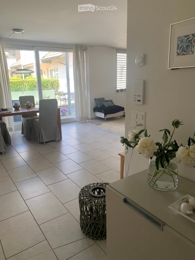 Open-plan living and dining area with tiled flooring, large windows offering a view, a dining table with chairs, a sofa, and a decorative vase with flowers on a side table.