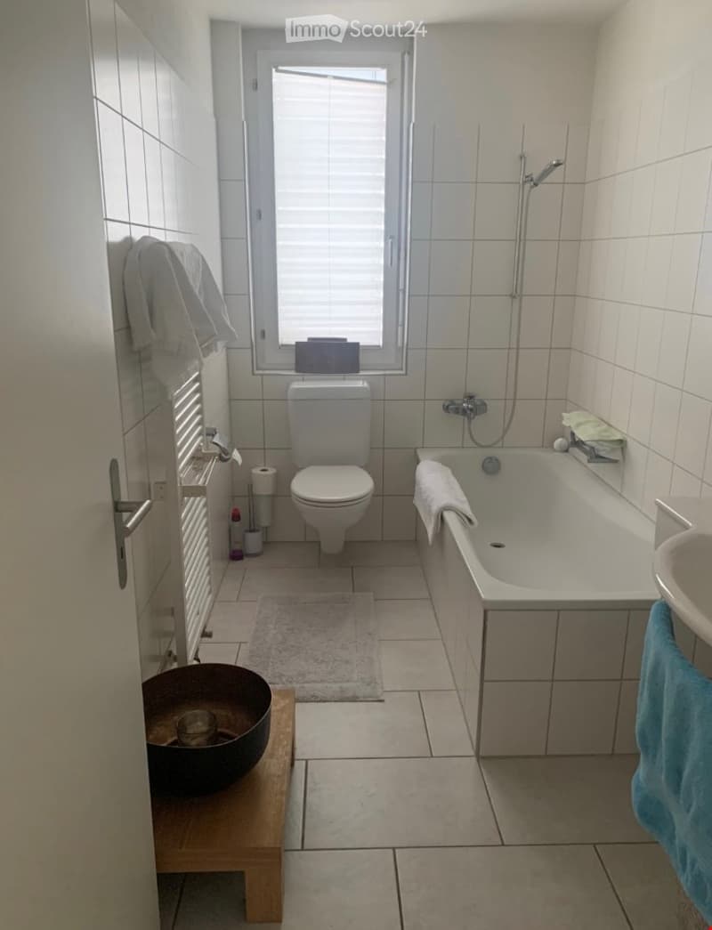 A modern bathroom with a toilet, bathtub, sink, and shower. The room features tiled walls and floor, a window with blinds, a towel rack, and a small wooden stool with a bowl on it.