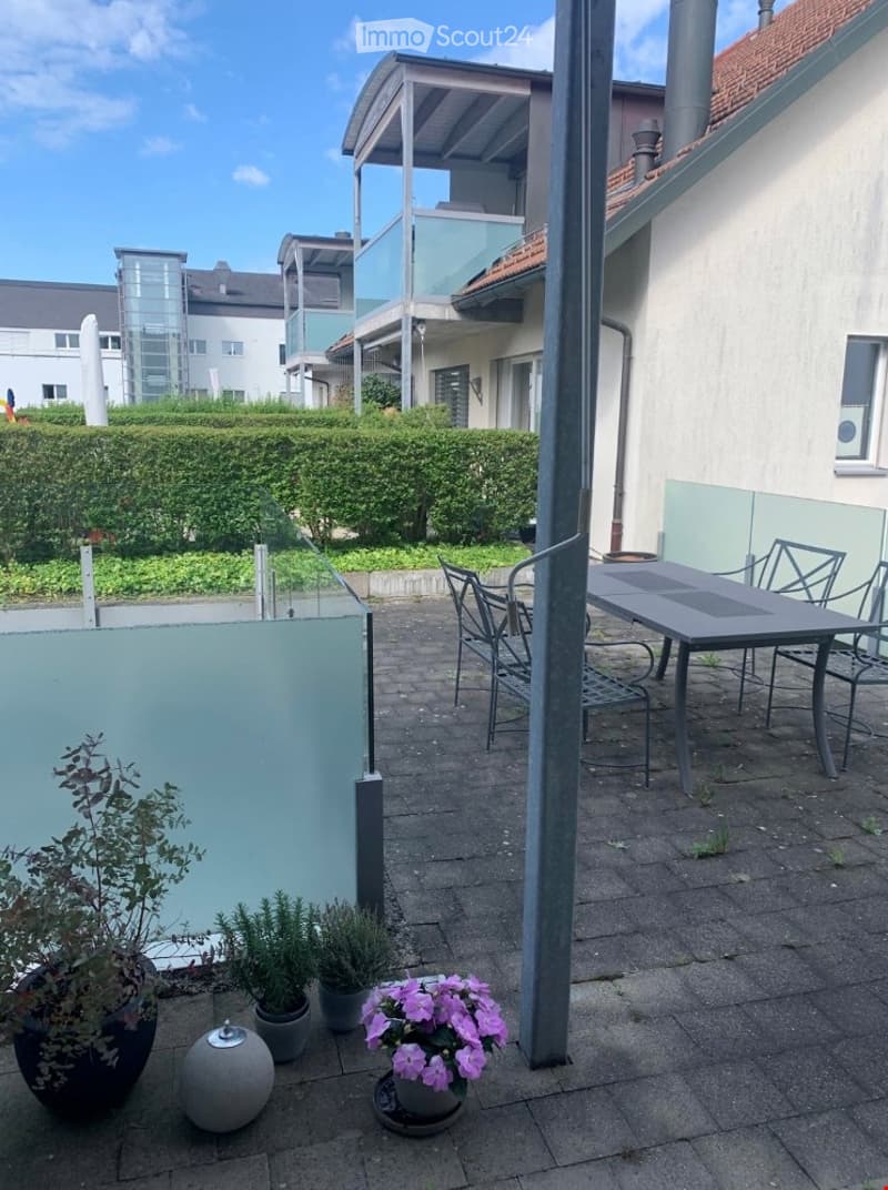 Balcony area with a table, chairs, and potted plants, surrounded by modern buildings and greenery.