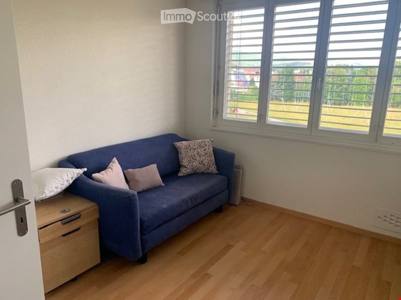 A small, modern living room with a blue sofa, decorative pillows, and a wooden side table. The room features wooden flooring and large windows with blinds, offering a view of greenery outside.