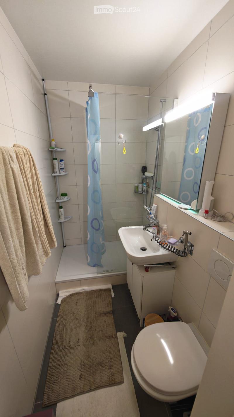 Modern bathroom with a bathtub, sink, toilet, mirror, and a towel hanging on the wall. A shower curtain is visible in the bathtub area, and a small shelf holds toiletries.