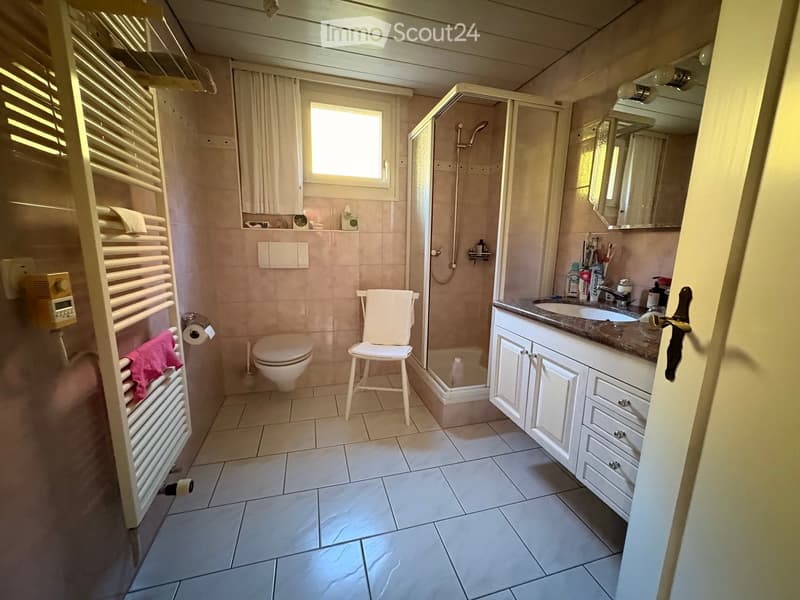 Bathroom with white tiled floor and walls, toilet, sink with mirror, shower, radiator, and a small stool.