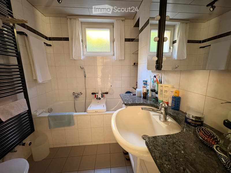 A well-lit bathroom with a white tiled wall, a bathtub, a sink with a faucet, a mirror, a towel rack with towels, and various toiletries on the countertop.
