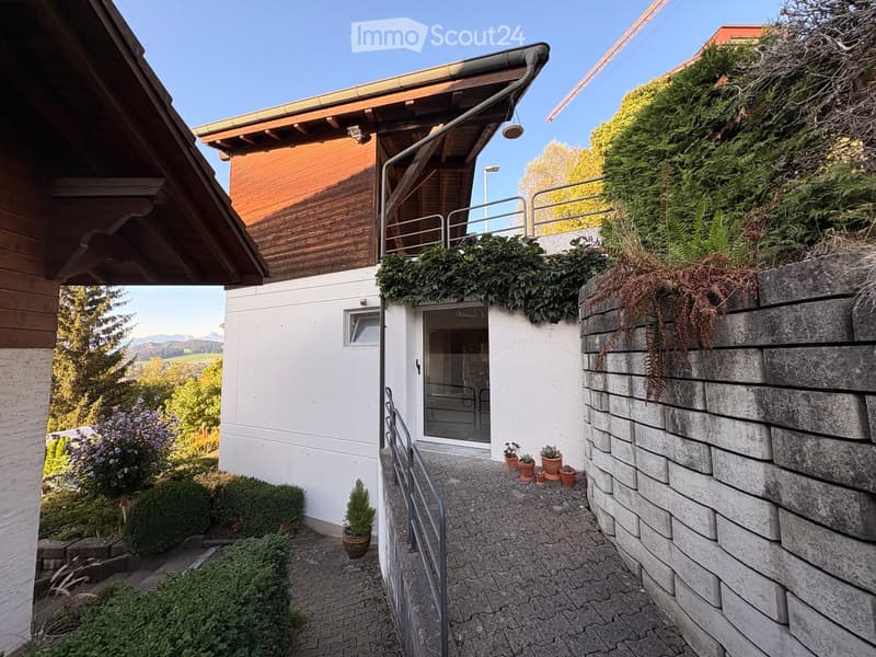 A serene outdoor area with a stone wall, potted plants, and a view of a lush green landscape and mountains in the background.