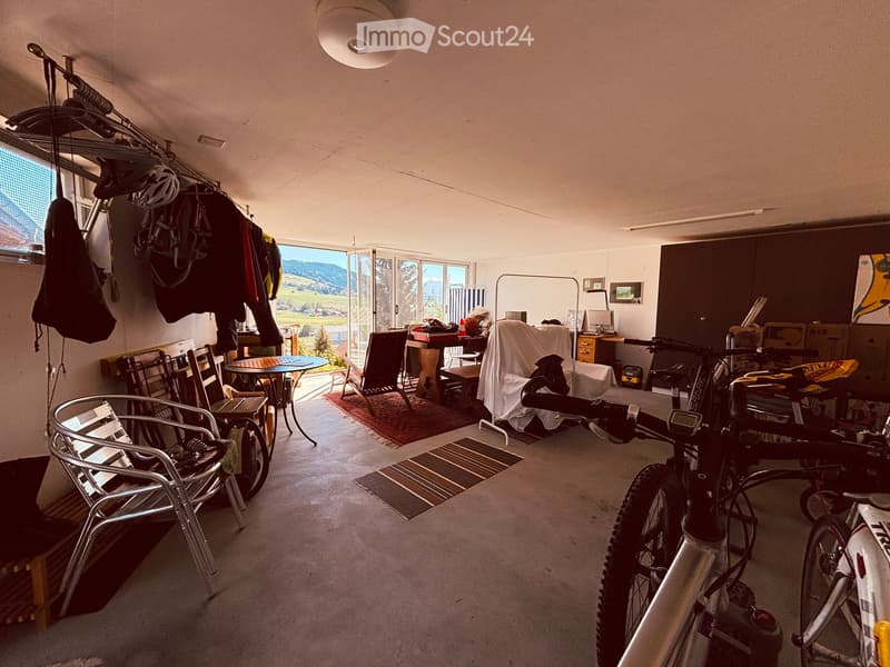A well-lit room with a view of the landscape, featuring a table and chairs, a bicycle, and a bench. The room includes a cabinet, a lamp, and a covered object on the floor.