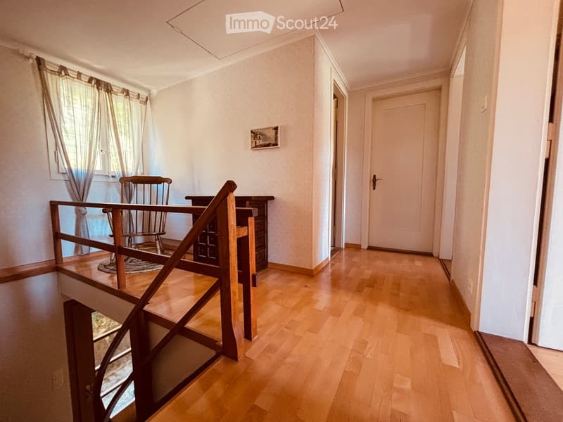 Spacious hallway with wooden flooring, a staircase leading to an upper level, a small table and chair in the corner, and a closed door at the end.