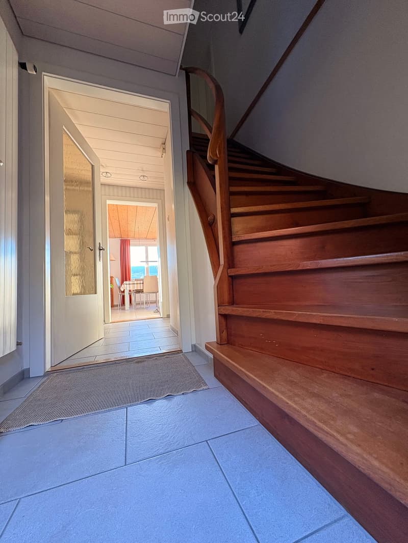 Hallway with wooden staircase, tiled floor, and open doorway leading to a dining area with a table and chairs.