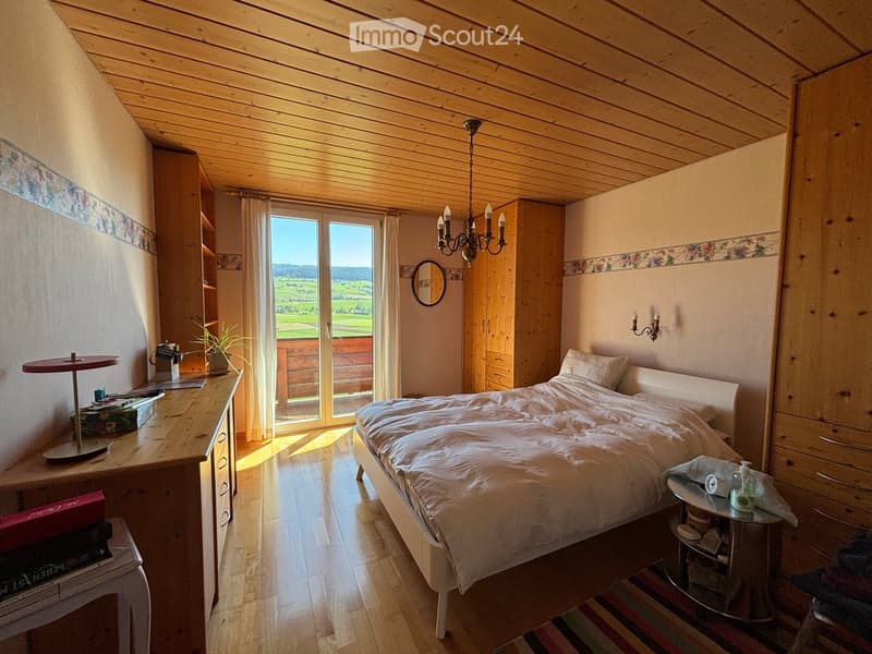 Cozy bedroom with wooden ceiling, large window offering a scenic view, bed with white bedding, nightstand, mirror, chandelier, and a small table with a plant.