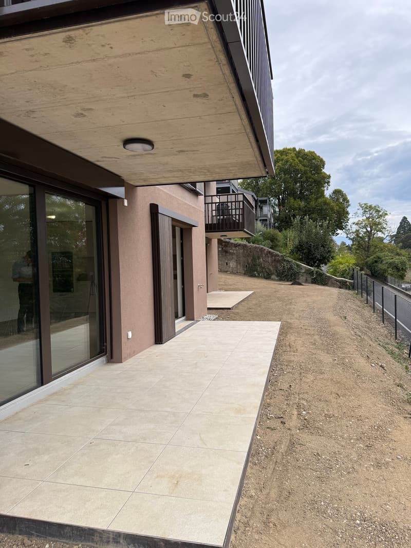 Modern house exterior with large glass windows, tiled patio, raised ground floor, and a gravel pathway leading to the entrance.