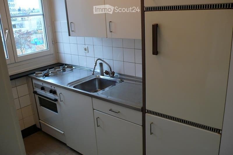 Compact kitchen with white cabinets, a four-burner stove, a sink with a faucet, and a refrigerator. The space features tiled walls and a window offering a view of the outside.