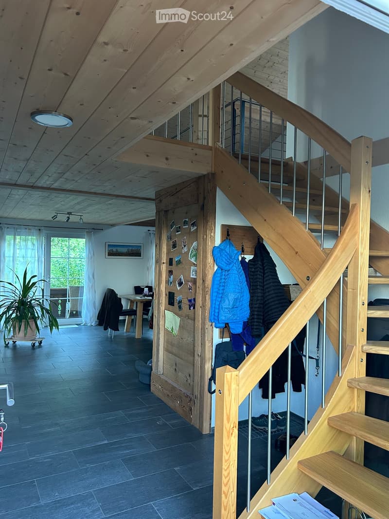 Bright hallway with wooden staircase, coat rack, and open living area visible through doorway. Features exposed wooden beams, modern lighting, and a potted plant near the entrance.