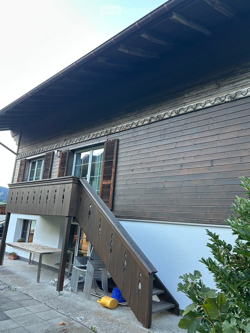 Two-story wooden house with a balcony, wooden shutters, and a staircase leading to the entrance.