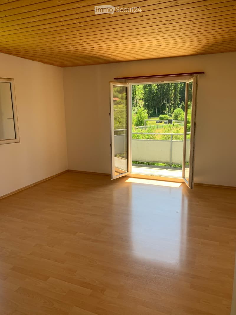 Empty room with wooden flooring, white walls, and a large open sliding door leading to a balcony with a view of greenery.