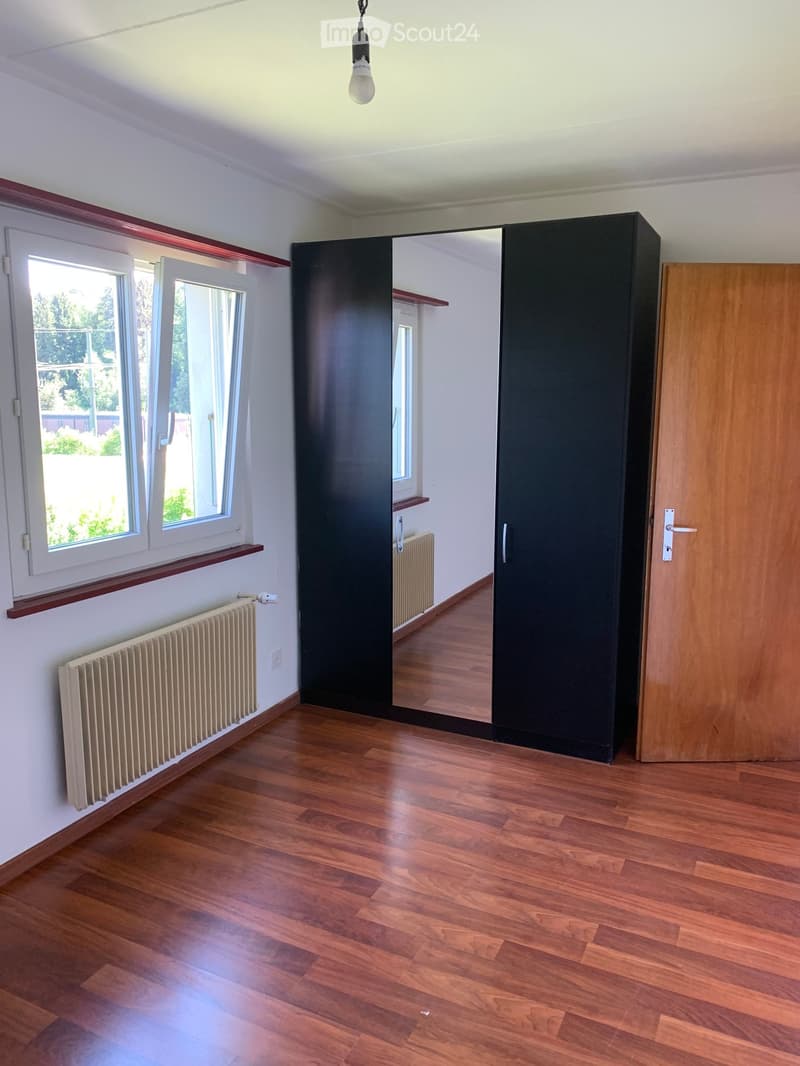 Empty room with wooden flooring, white walls, large windows, and a black wardrobe. No furniture or equipment present.