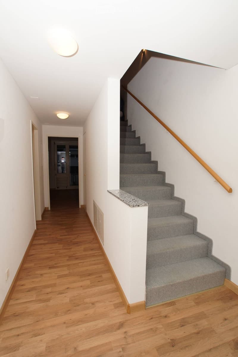 {"visible_features": [], "type": "Room", "description": "Interior hallway with white walls, wooden flooring, gray carpeted stairs, and a wooden handrail."}