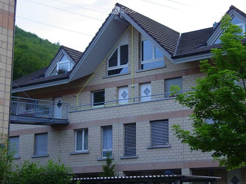 Three story apartment building, brick facade, brown roof, balconies on second and third floor, greenery in the foreground