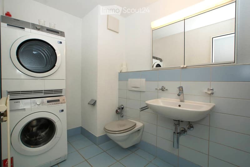 Small bathroom with white walls, blue tiles, white sink, shower, toilet, and a washing machine on the left side.