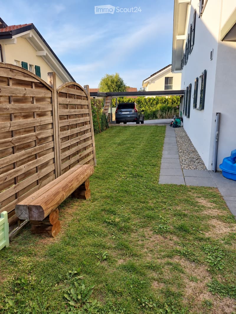 A well-maintained garden area with a wooden fence, a paved pathway, and a small seating area with a wooden bench. The area is bordered by a house and includes a driveway leading to a parked car.