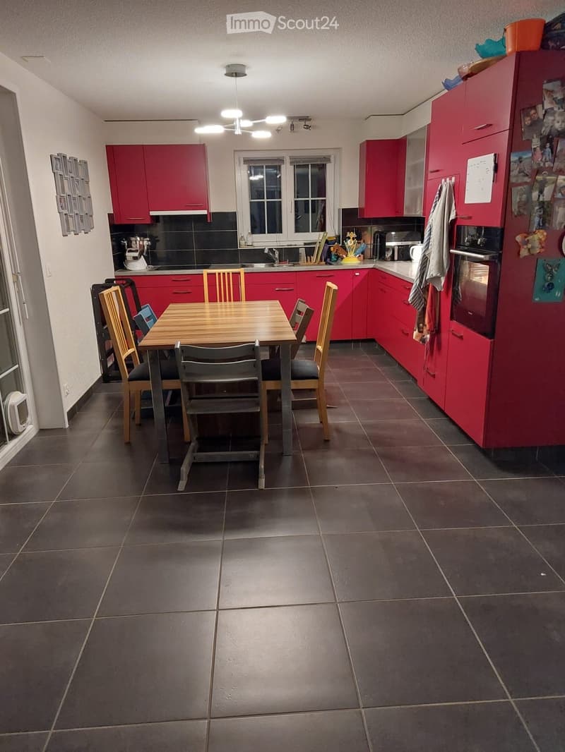 Modern kitchen with red cabinets, a wooden dining table with four chairs, a refrigerator, and various appliances.