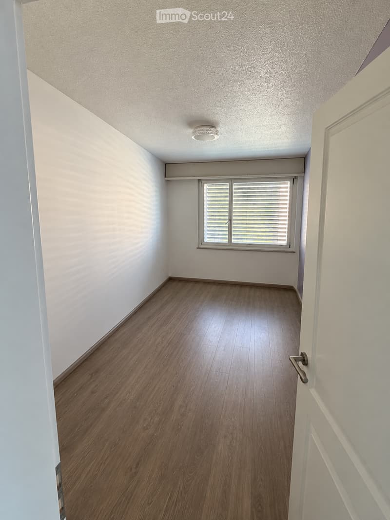 Empty room with wooden flooring, white walls, and a single window with blinds. The room has a ceiling light fixture and a door with a handle visible on the right side.