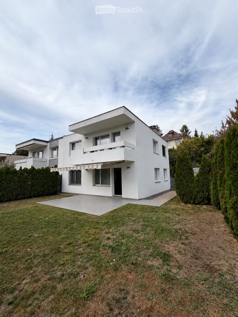 Modern two-story white house with flat roof, multiple windows, balconies, and a covered entrance. The house is surrounded by a well-maintained lawn, trimmed hedges, and tall evergreen trees.