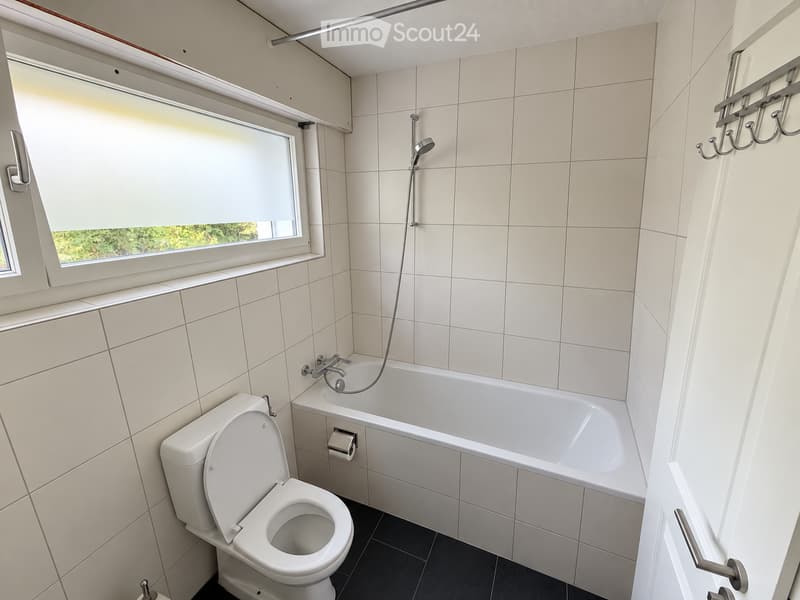 Modern bathroom with white tiled walls and floor, featuring a toilet, bathtub, shower hose, and a window with blinds.