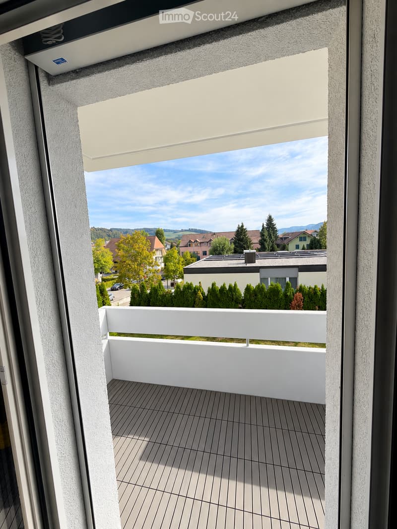 Balcony with wooden decking, white railing, and a view of a suburban neighborhood with houses, trees, and mountains in the background.