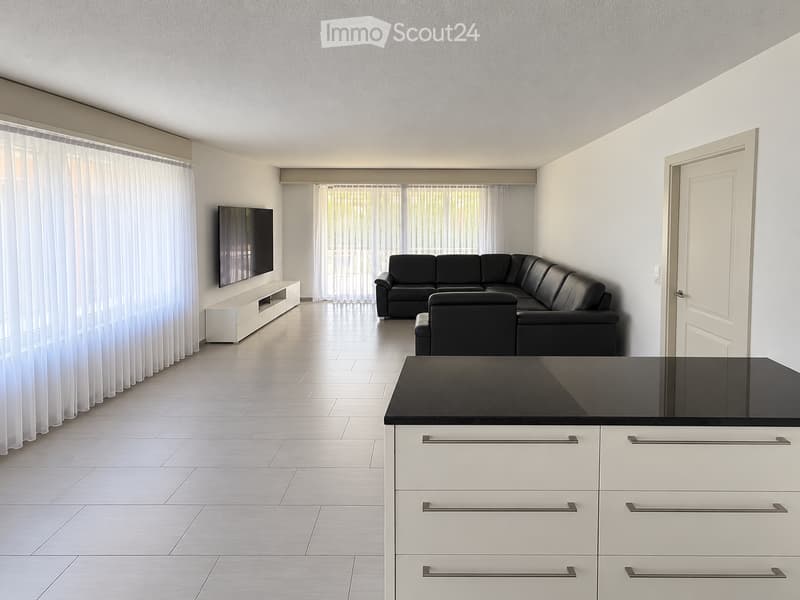 Modern living and dining area with a black sectional sofa, flat-screen TV on a white stand, and a white kitchen island with black countertop.