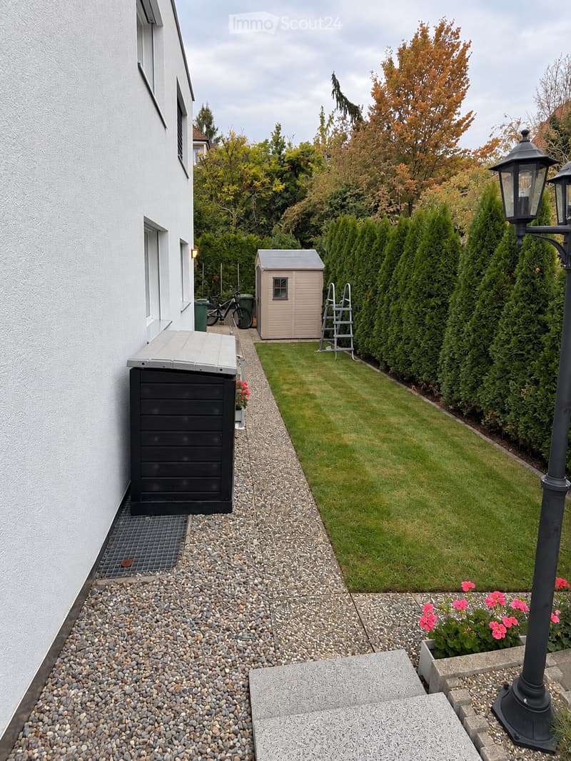 Well-maintained garden with a neatly trimmed lawn, gravel pathway, small shed, bicycle, and decorative plants near the entrance.