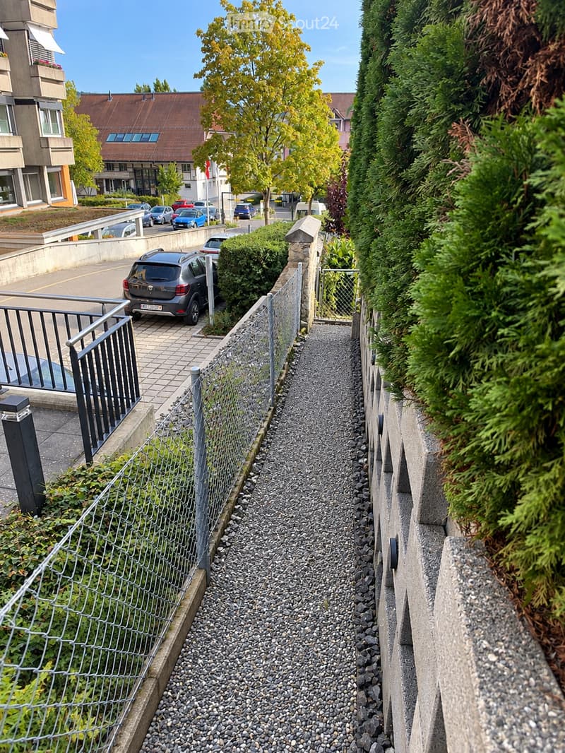 A narrow, elevated pathway lined with a metal fence, surrounded by greenery and leading to a parking area with several cars.