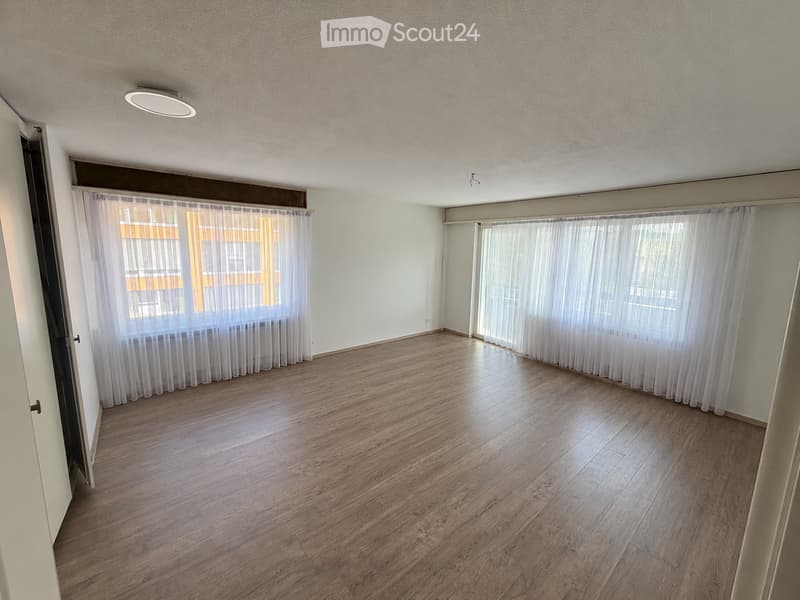 Empty room with wooden flooring, white walls, large windows with sheer curtains, and a ceiling light fixture.
