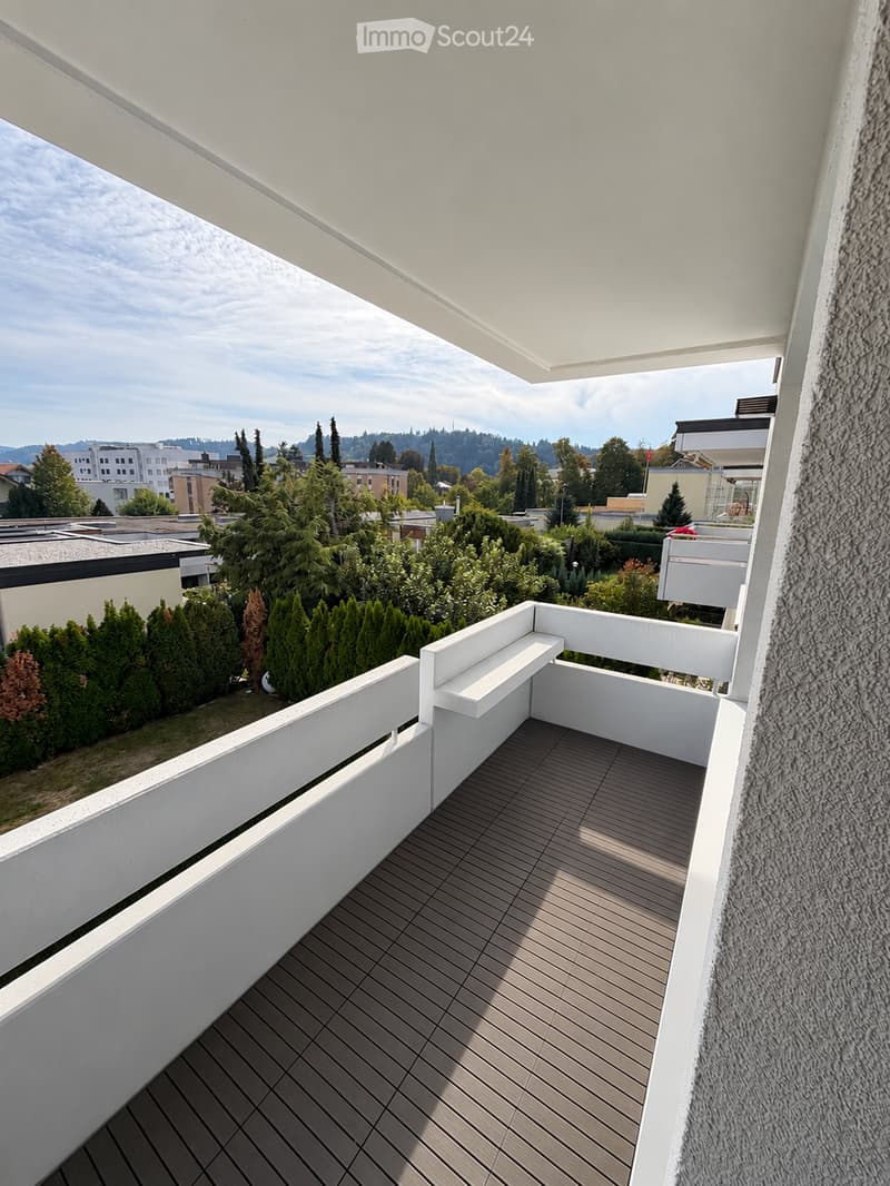 Modern balcony with wooden decking, white railings, and a bench, overlooking a landscaped area with trees and buildings.