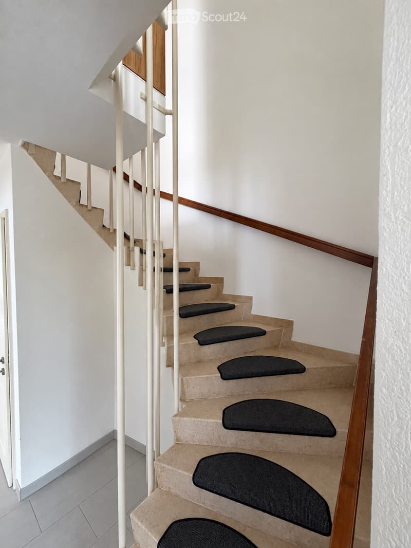 Spiral staircase with beige stone steps and black rubber treads, white walls, wooden handrail, and a white door visible on the left side.