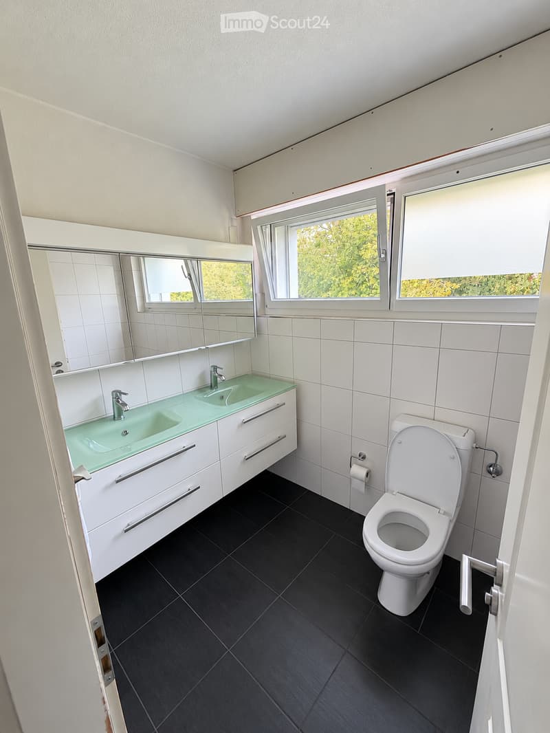 Modern bathroom with white tiled walls, black tiled floor, double sink with glass countertop, toilet, and large mirror above the sink. Windows provide natural light.