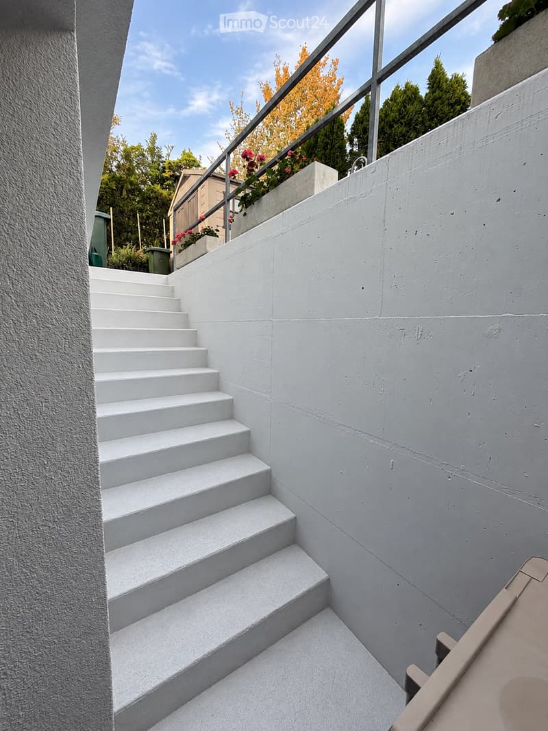 White concrete staircase leading up to a raised platform with a metal railing, surrounded by greenery and a small planter with flowers.