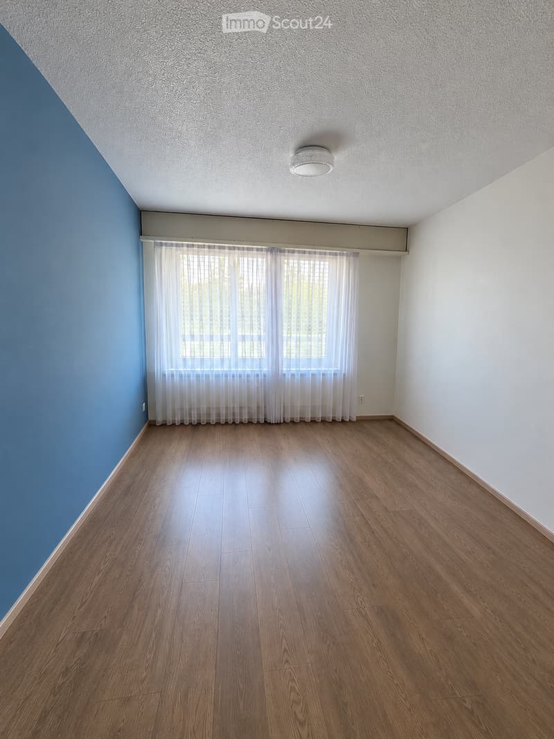 Empty room with wooden flooring, white walls, and a large window with white curtains. The ceiling has a light fixture, and the walls are painted in blue and white.