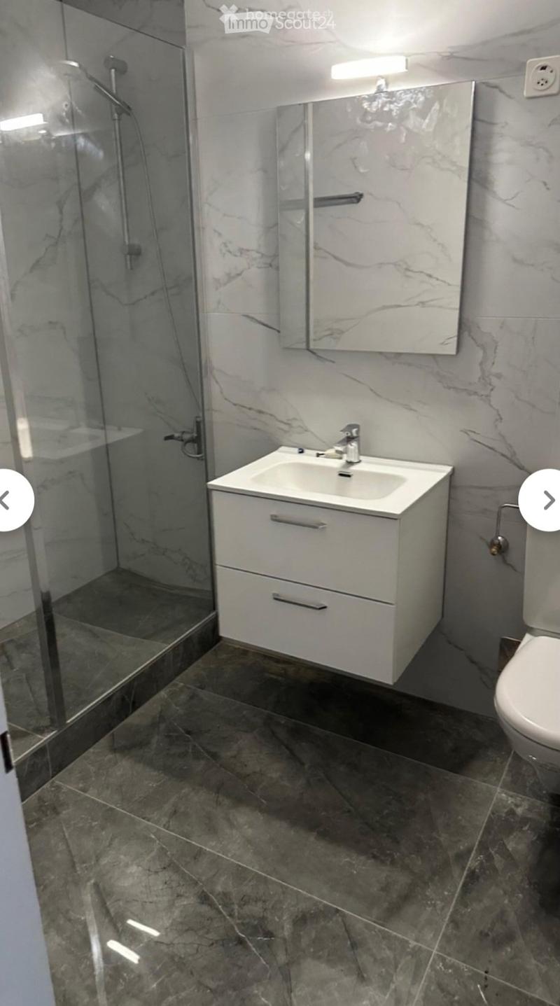 Modern bathroom with marble walls, dark tiled floor, white sink with two drawers, mirror, and glass-enclosed shower area.