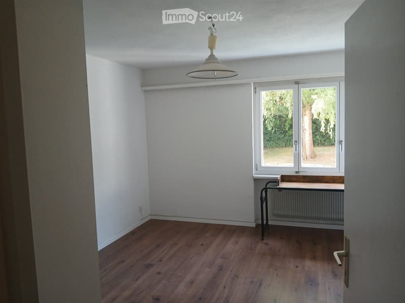 Empty room with wooden flooring, white walls, large window, and a simple hanging light fixture.