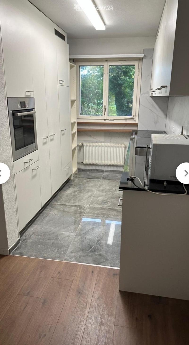 Empty kitchen with white cabinets, a built-in oven, and a window overlooking greenery. The floor is a mix of dark tiles and light wood, and the space is well-lit with natural light.
