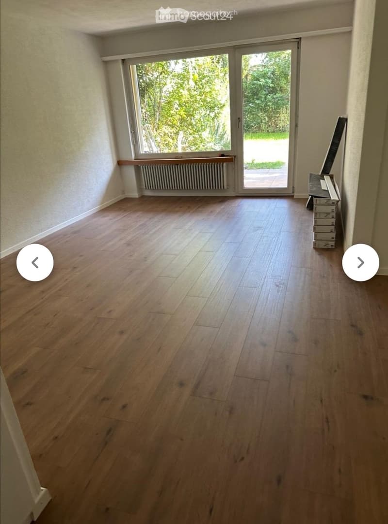 Empty room with wooden flooring, large sliding glass door leading to outdoor area, radiator under window, and a stack of boxes in the corner.