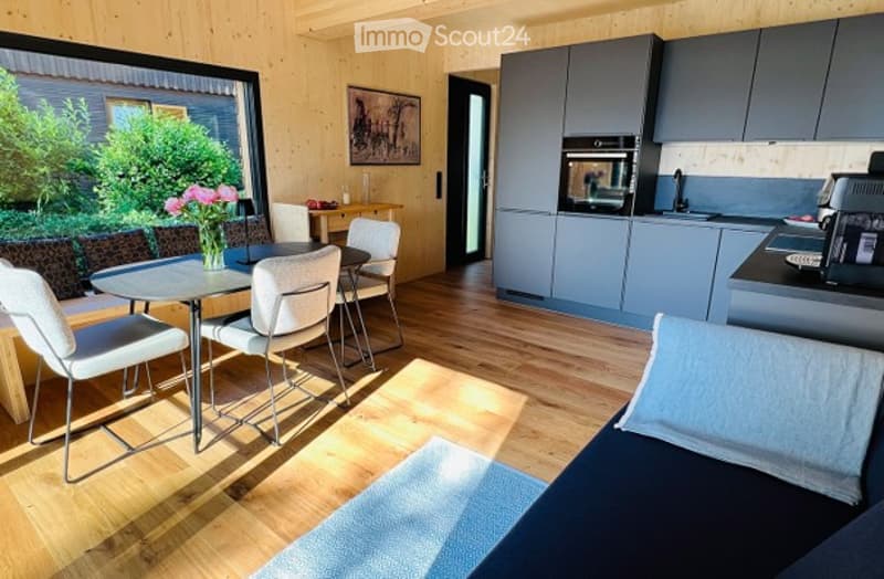 Bright, modern living and dining area with wooden flooring, large windows offering a view of greenery, a sofa, coffee table, dining table with chairs, and a modern kitchen area with cabinets, fridge, microwave, and oven.