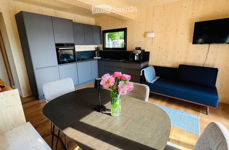 A modern, open-plan living and dining area with grey kitchen cabinets, a blue sofa, a dining table with chairs, and a vase of pink flowers on the table. The room features wooden walls and a television mounted on the wall.