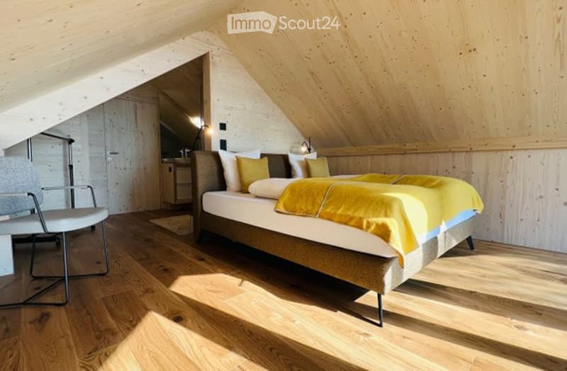 Cozy attic bedroom with wooden walls and floors, a large bed with yellow bedding, a modern chair, and a nightstand with a lamp.