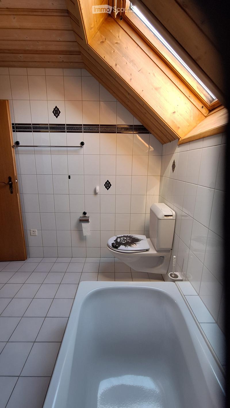 Modern bathroom with a bathtub, toilet, and a toilet paper holder. The walls are tiled with white tiles and decorative black accents. A wooden door is visible on the left side.