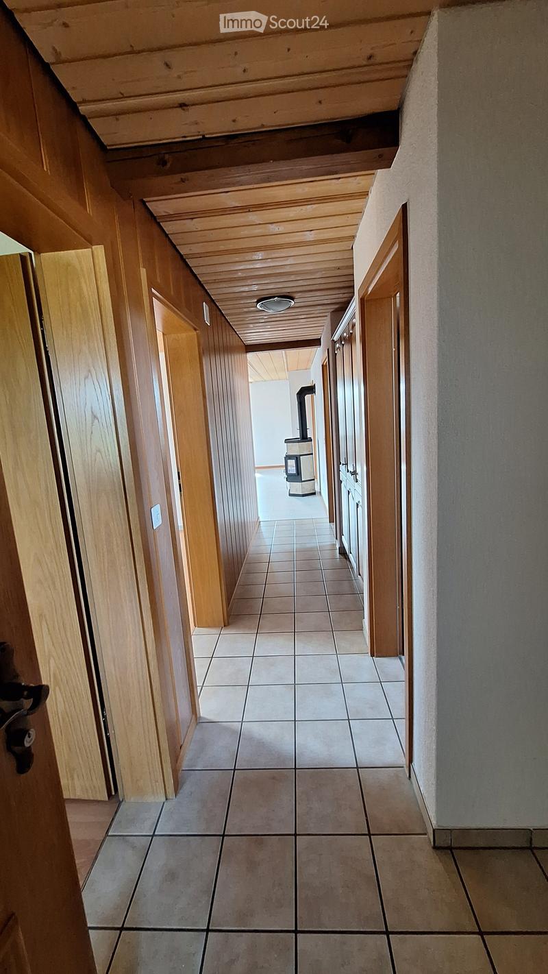 A long, narrow hallway with tiled flooring, wooden paneling on one side, and several doors leading to rooms. The ceiling has exposed wooden beams and a central light fixture.